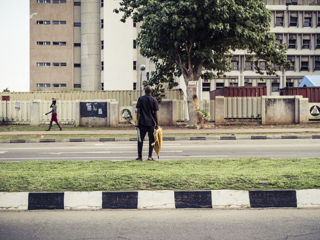 A person holding umbrellas stands by the roadside, with buildings and a tree visible in the background.